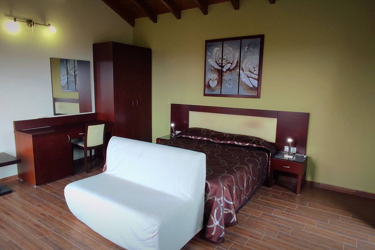 A bedroom with a bed, bedside table, nightstand, desk, chair, and artwork on the wall.