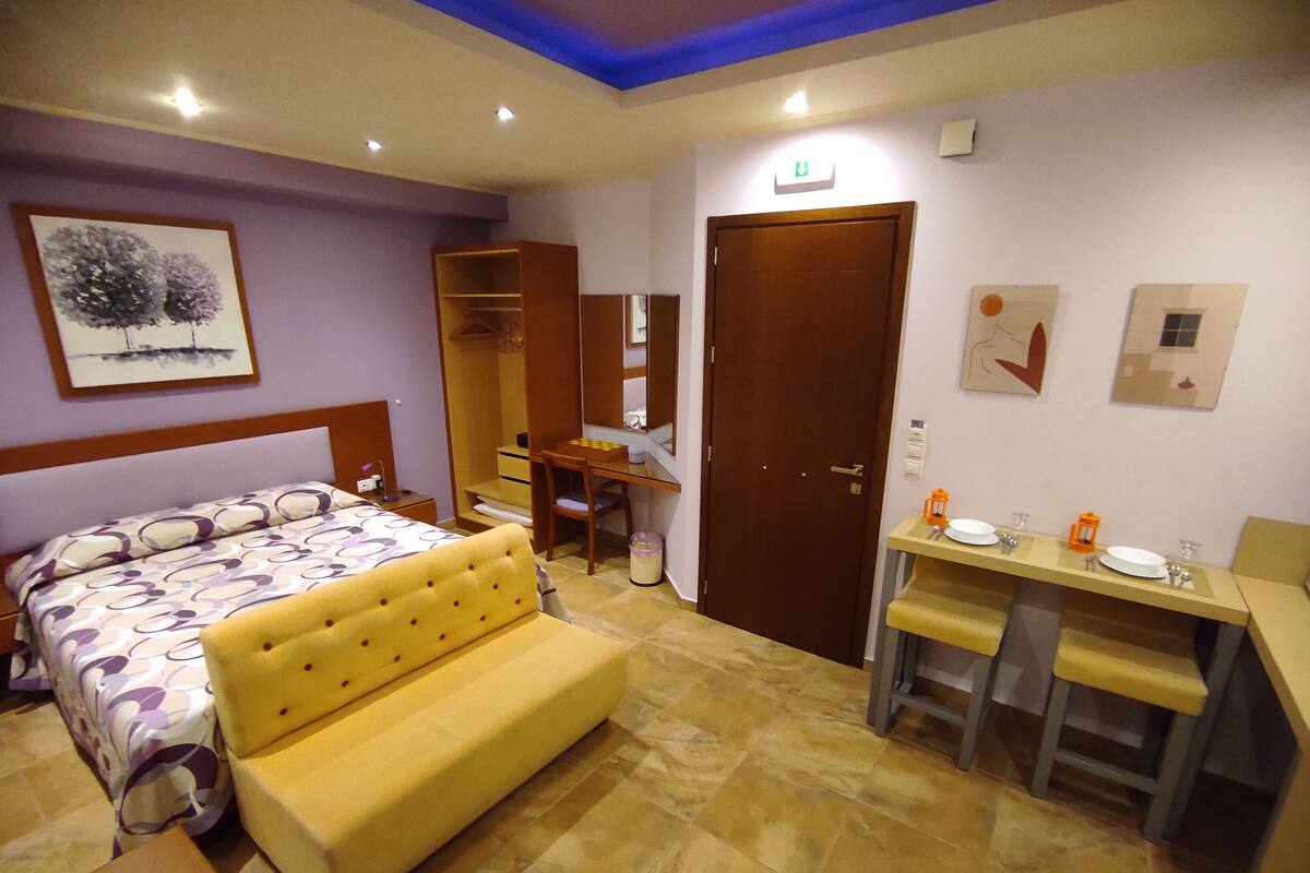A bedroom with a bed, a sofa, a desk with a chair, a wardrobe, and a bathroom with a sink and mirror.