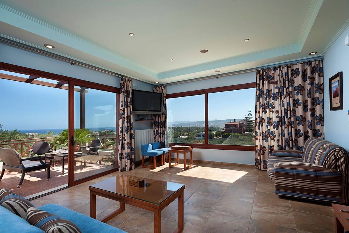 A spacious living room with a large TV, comfortable seating, and a view of the sea.