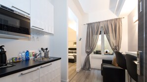 A modern kitchen with a built-in oven, a sink, and a countertop with various items. There is a window with curtains, a bed in the adjacent room, and a small seating area with a chair. - Brunelleschi Holidays Hostel (Rome)