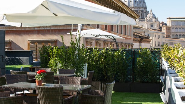 An outdoor patio with wicker furniture and white umbrellas, a well-maintained lawn, and a view of a historic building with a dome. - Brunelleschi Holidays Hostel (Rome)