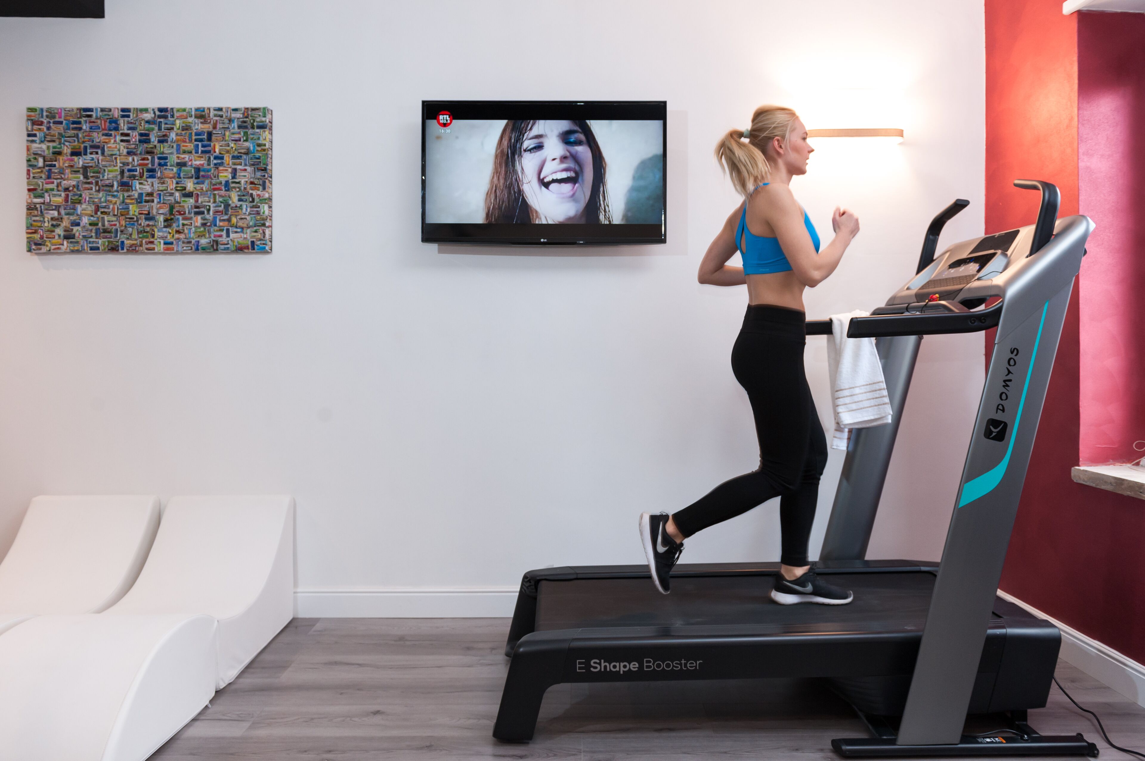 A woman running on a treadmill in a room with a television and a wall art piece.