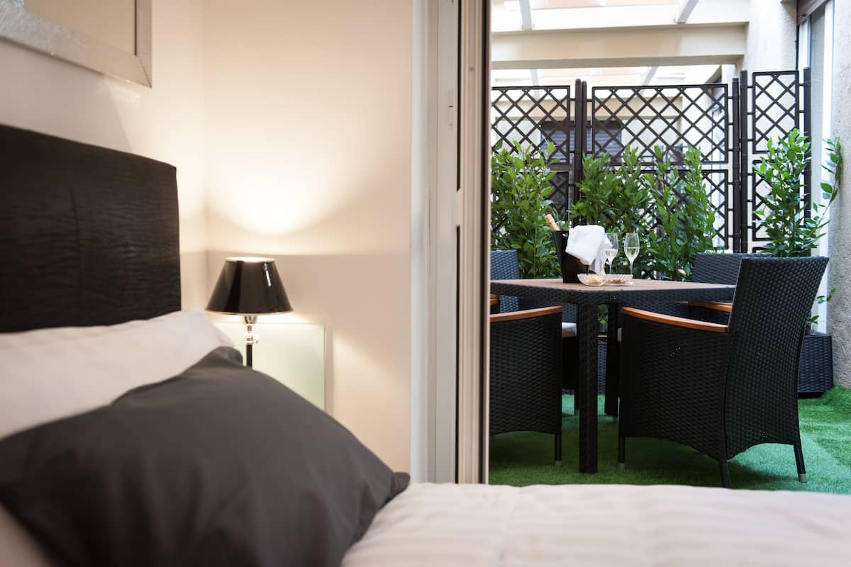 Ein Hotelzimmer mit Bett, Nachttisch, Lampe und Blick auf einen Außenbereich mit Tisch, Stühlen und Grünpflanzen.