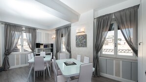 A dining area with a glass table and white chairs, a kitchen area with cabinets, and large windows with curtains. - Brunelleschi Holidays Hostel (Rome)