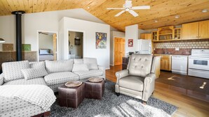 A cozy living room with a sofa, armchair, and ottomans, a kitchen with wooden cabinets, and a ceiling fan.