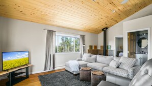 A modern living room with a wooden ceiling, a large window with curtains, a grey sofa, a leather armchair, and a flat-screen TV on a wooden stand.