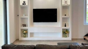 A modern living room with a large flat-screen TV mounted on a white wall, surrounded by built-in shelves and a dark-colored sofa. - Magnifique Maison Avec Jacuzzi (Marseille)