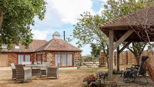 A rustic outdoor seating area with wicker furniture, a covered patio, and a brick building with a chimney in the background. - Baa Lamb Cottage (Romney Marsh)