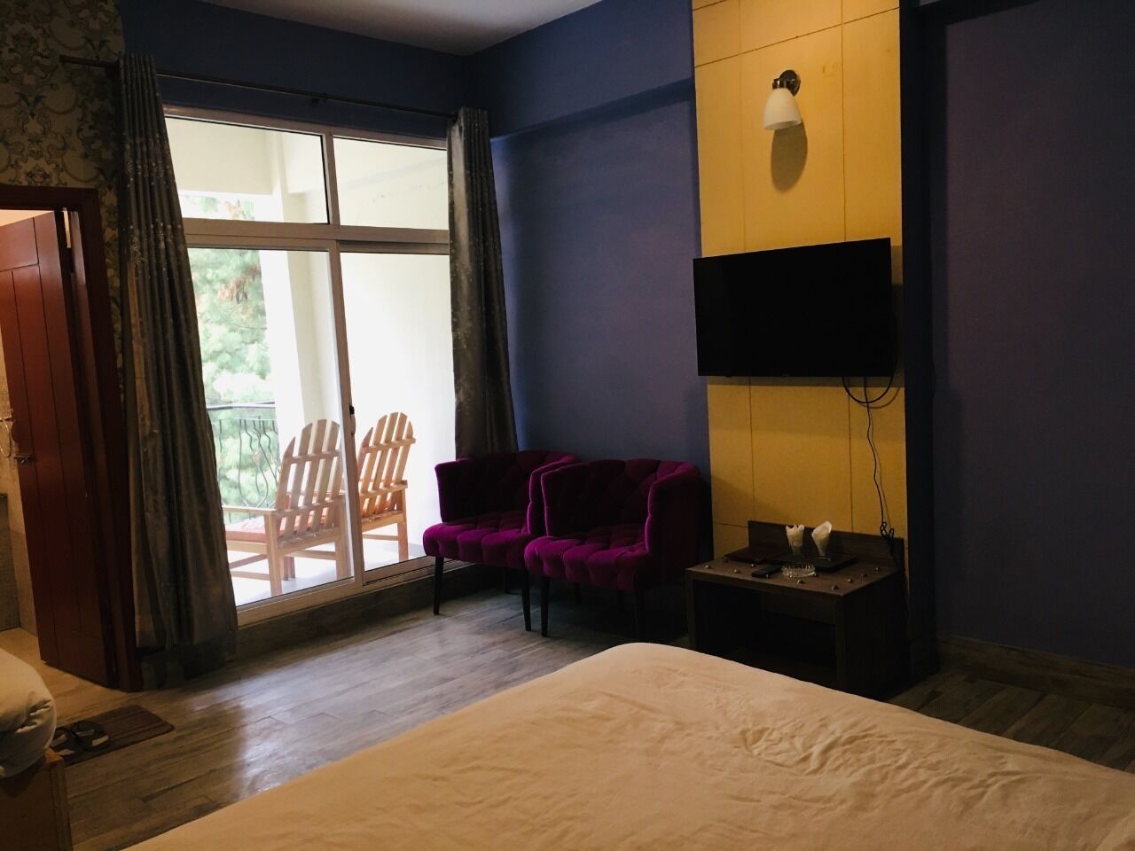 A hotel room with a large bed, a TV on the wall, a desk with a chair, and a window with a view of outdoor seating.