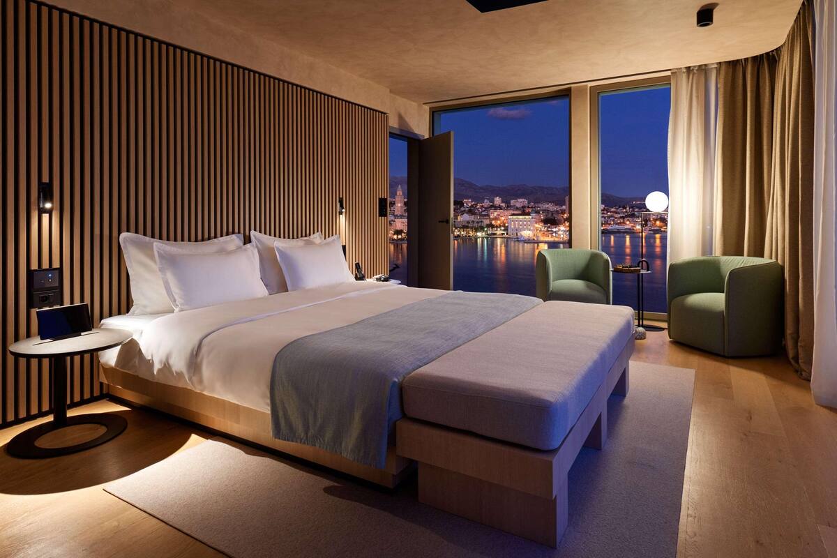 A modern hotel room with a large bed, a small table, a chair, and a view of a cityscape at night.