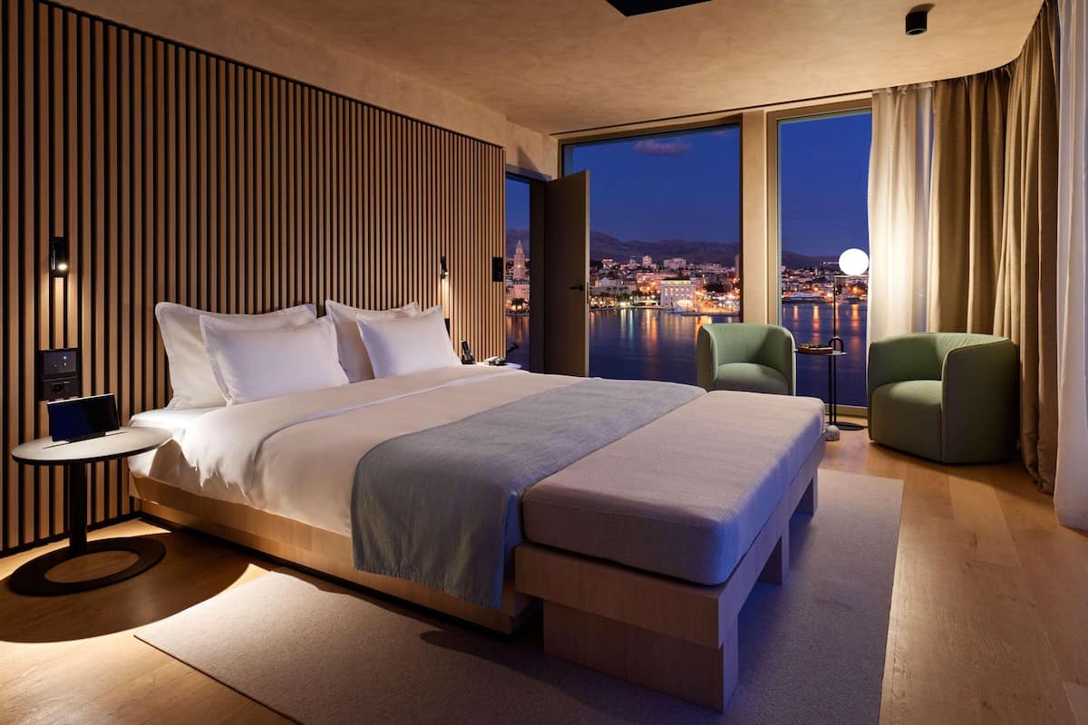 A modern hotel room with a large bed, a small table, a chair, and a view of a cityscape at night.