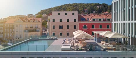 A rooftop pool area with lounge chairs and umbrellas, surrounded by buildings with red and white facades.