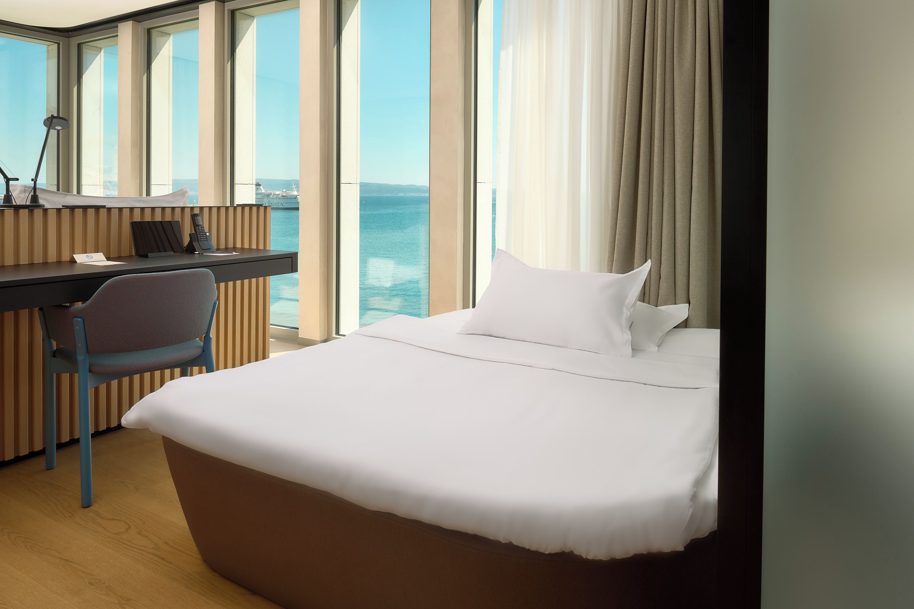 A hotel room with a bed, a desk, a chair, and a view of the sea.