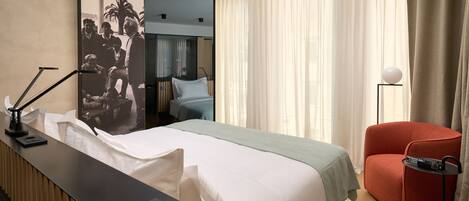 A hotel room with a bed, a desk with a lamp, a red chair, a remote control, and a large poster on the wall.