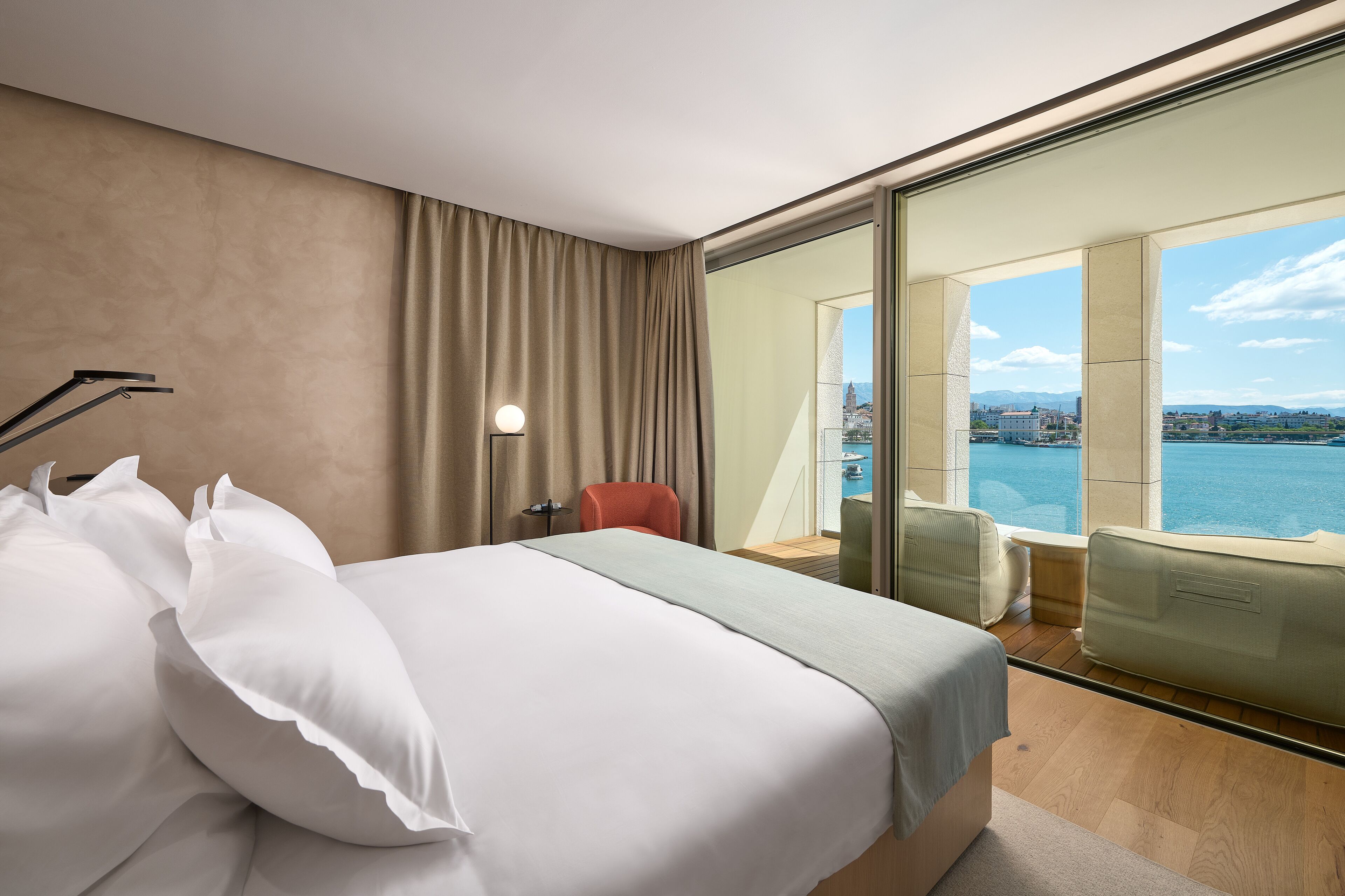 A modern hotel room with a large bed, a red chair, a desk, and a view of the city and water.
