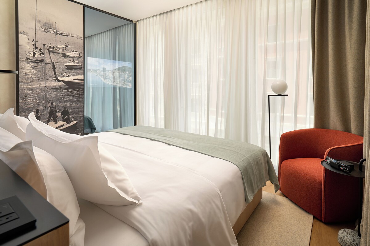 A hotel room with a large bed, a red armchair, a floor lamp, and a painting on the wall.
