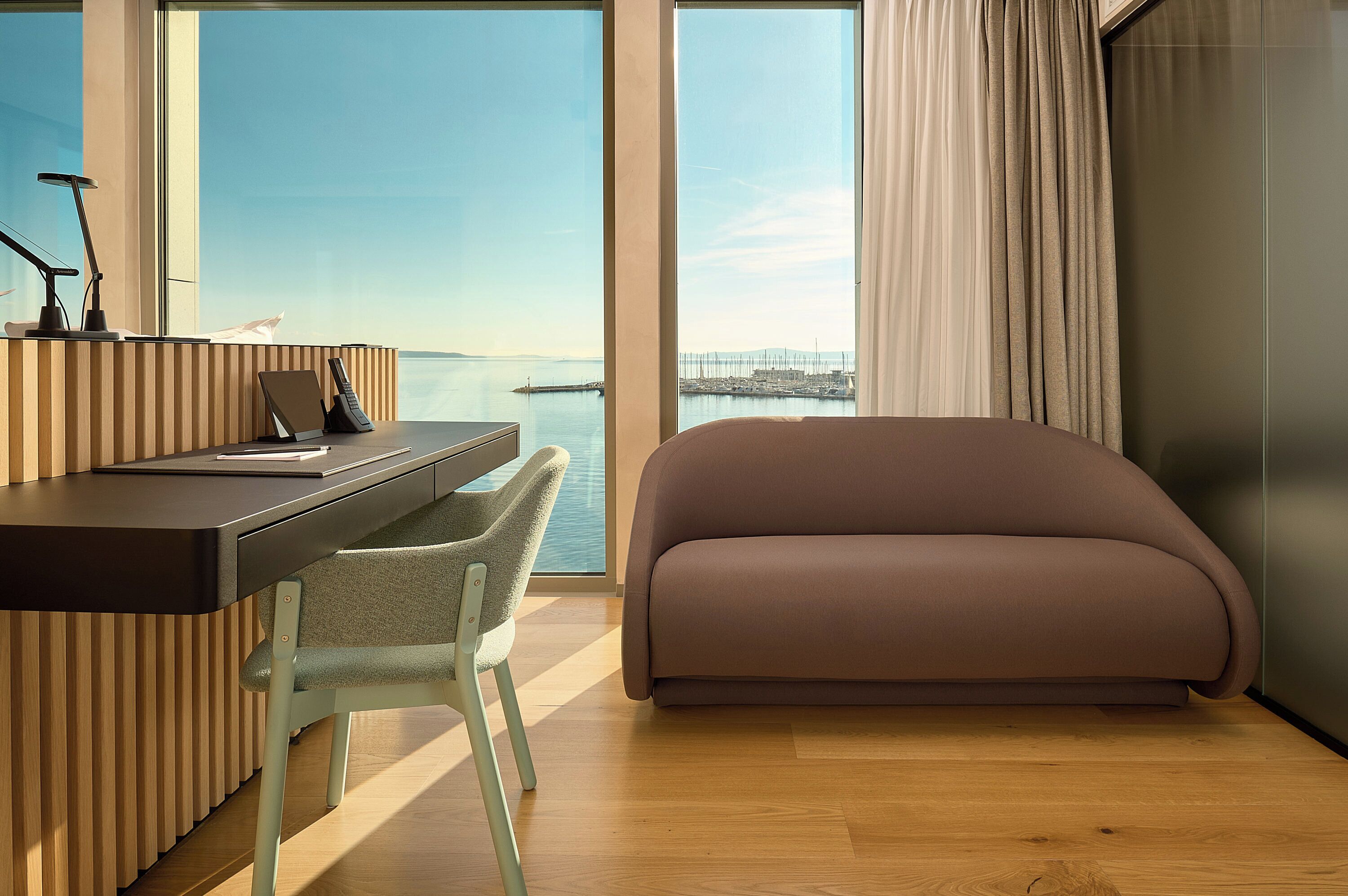 Una habitación moderna con escritorio, silla y sofá, ventanas amplias con vista al mar y a un puente, y suelo de madera.
