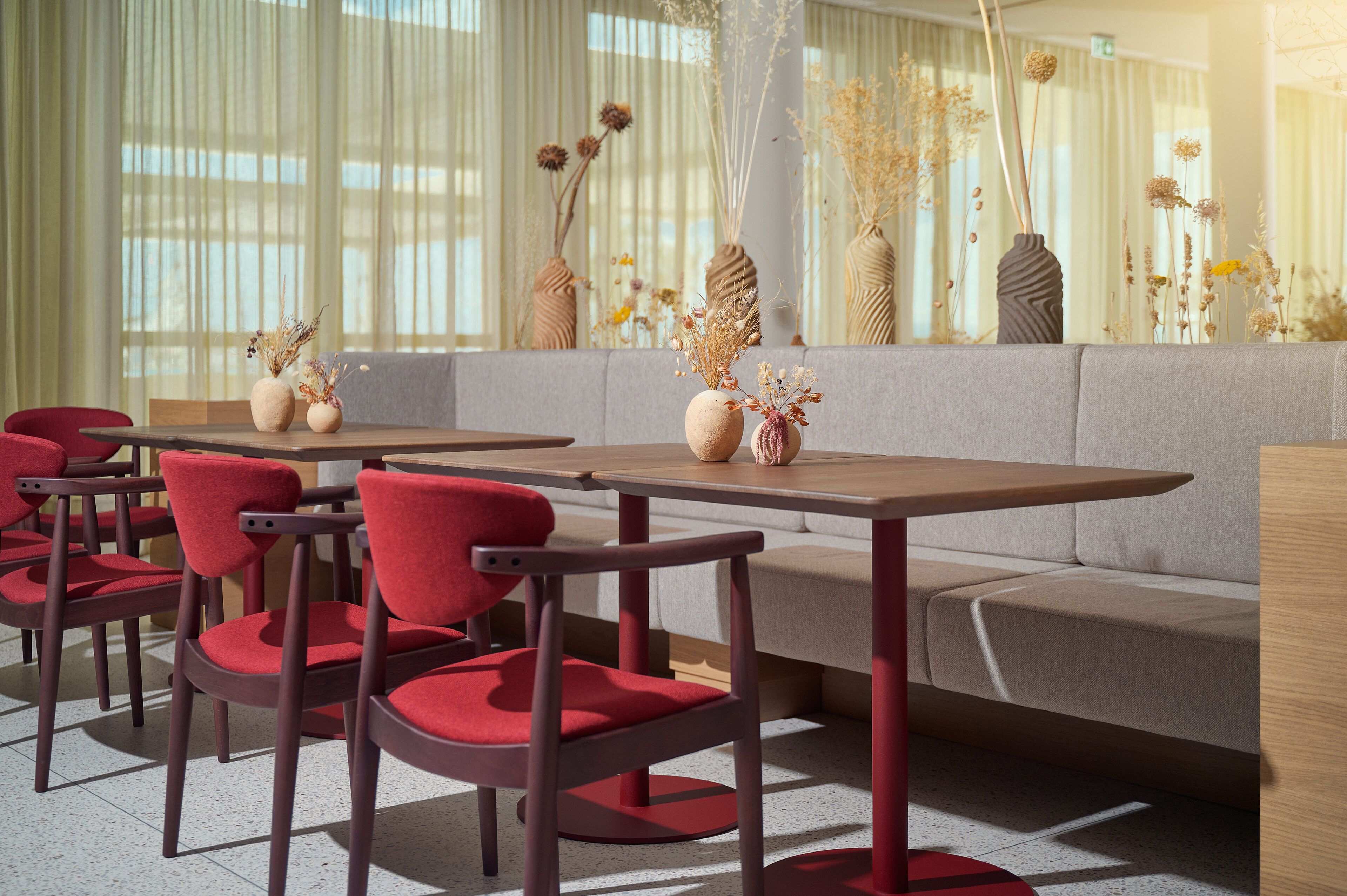 Un comedor moderno con sillas rojas, un sofá gris y varios jarrones con flores secas sobre una mesa.