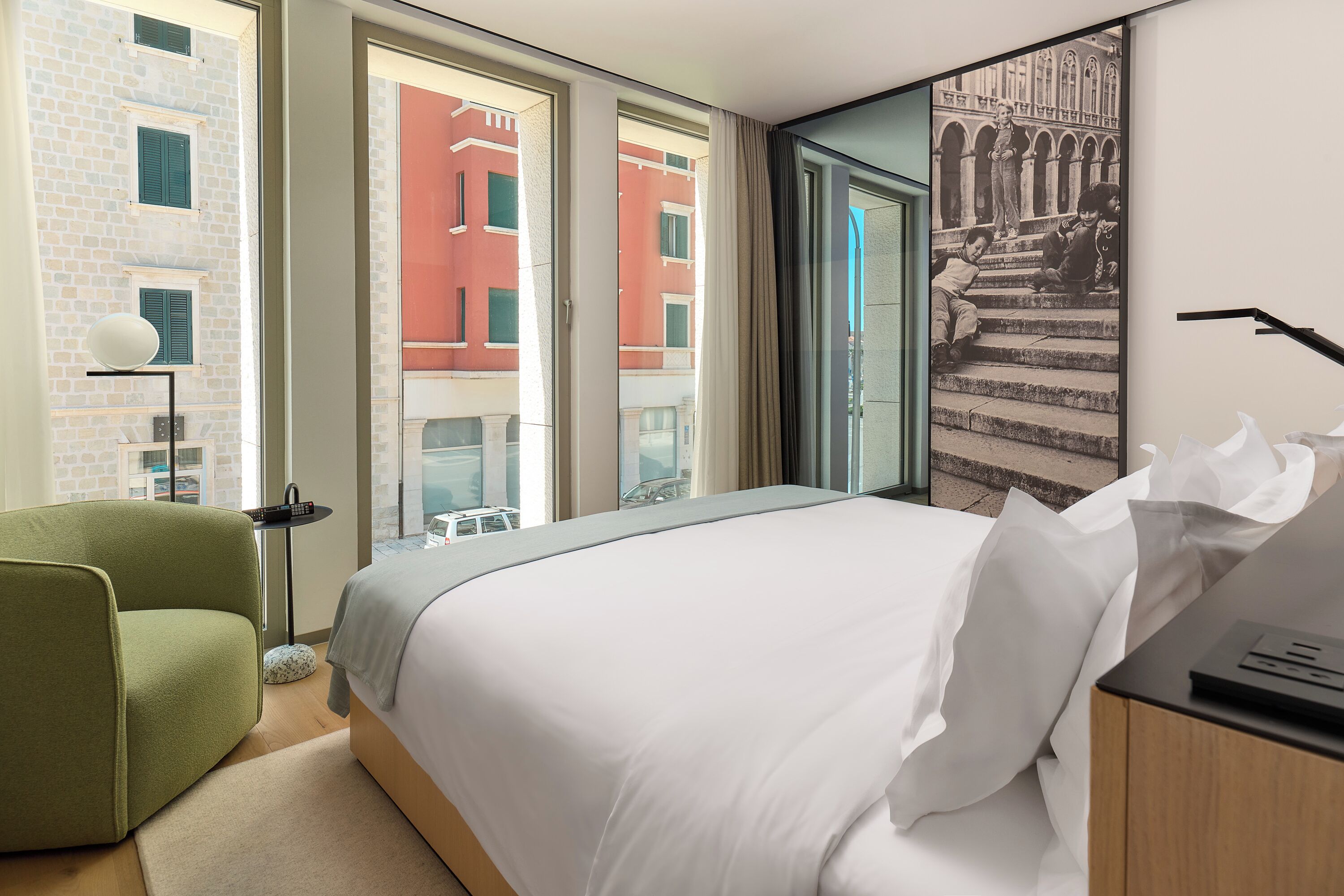 A hotel room with a large bed, a green armchair, a small table with a lamp, and a window with a view of buildings.