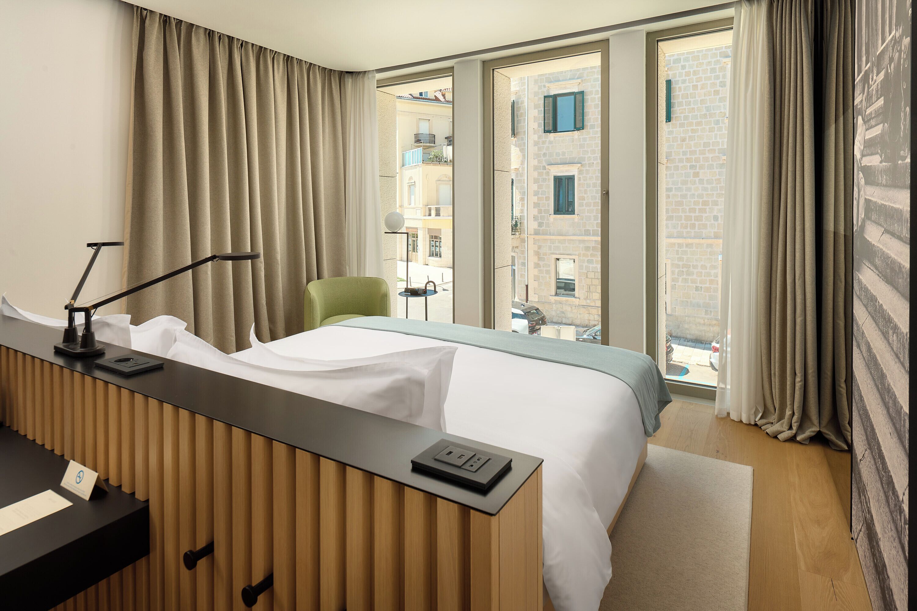 A hotel room with a bed, a desk with a lamp, a chair, and a view of buildings through the window.