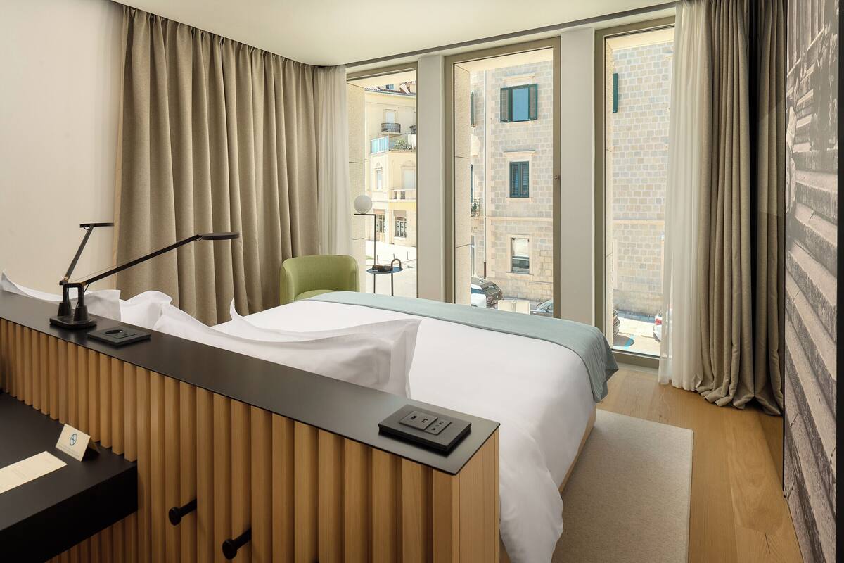 A hotel room with a bed, a desk with a lamp, a chair, and a view of buildings through the window.
