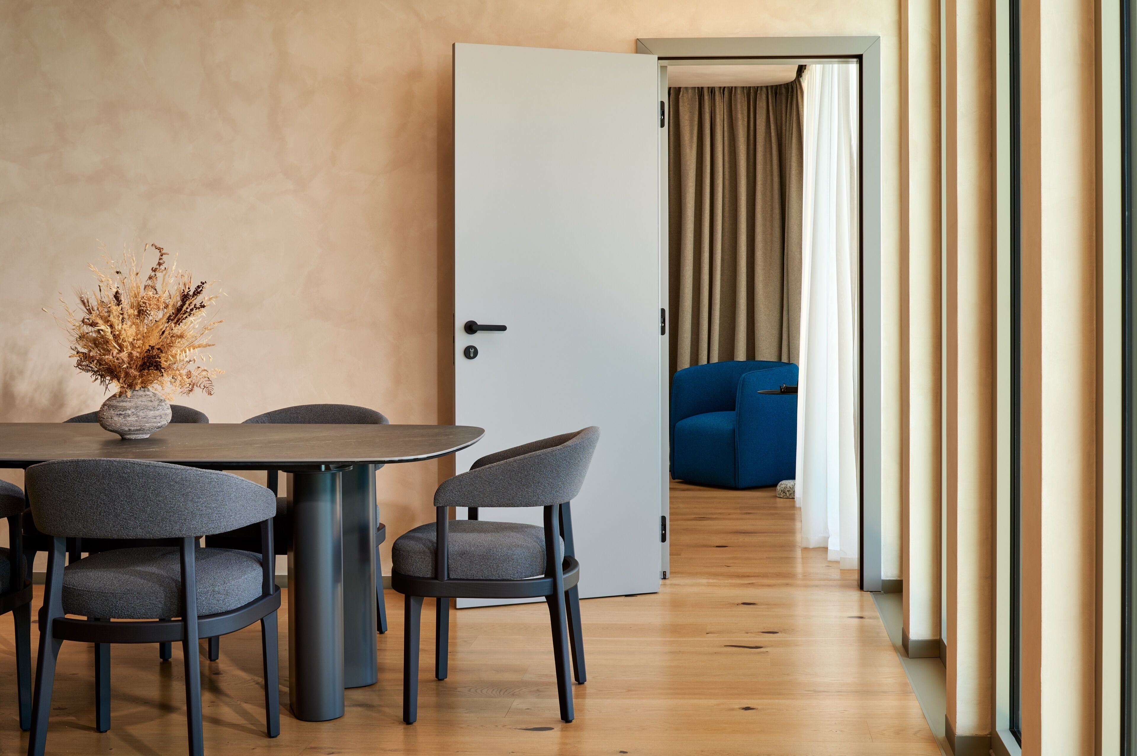 Une pièce moderne comprenant une table à manger, des chaises, un vase avec des fleurs séchées et une porte donnant sur une autre pièce où se trouve un canapé bleu.