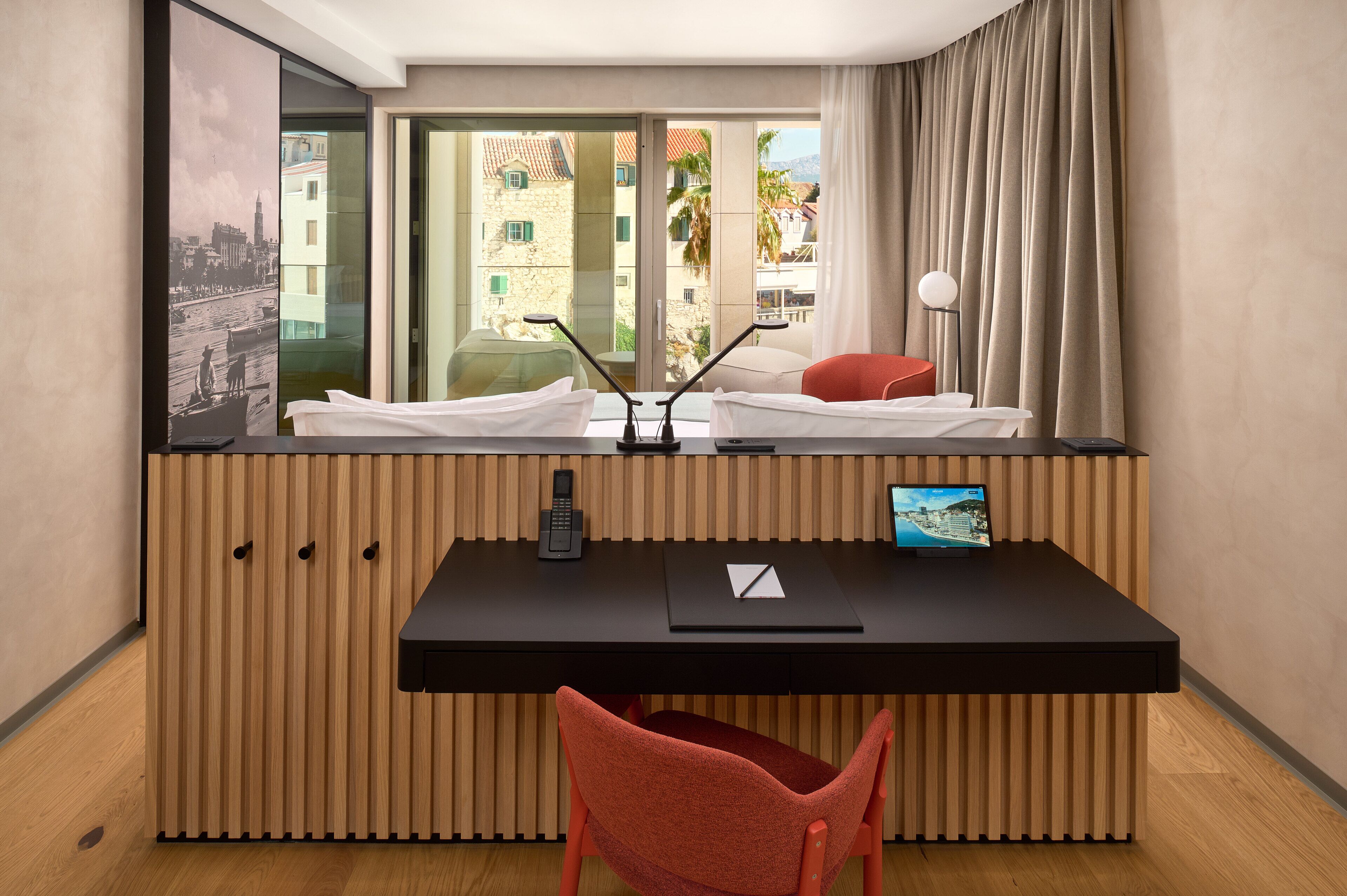 Una habitación de hotel moderna con un escritorio de madera, una silla roja, un ventanal con vistas a edificios y palmeras, y una pintura de paisaje urbano.