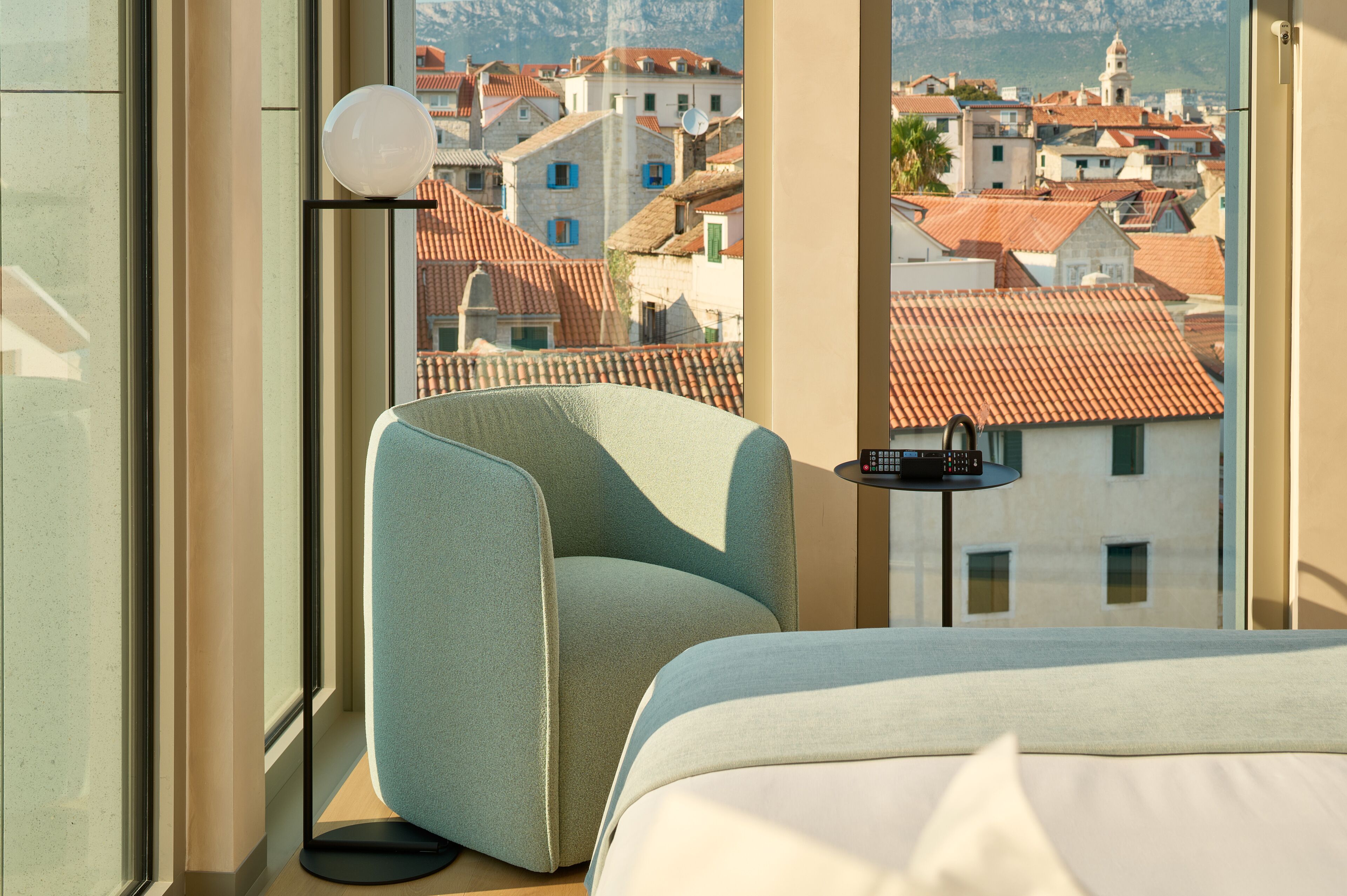 Una camera d'albergo con un letto, una sedia, un tavolo e vista su un paesaggio urbano con edifici dai tetti rossi.