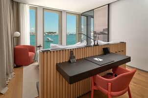 A modern hotel room with a desk, a red chair, a large window with a view of the sea and boats, and a wooden panel wall.