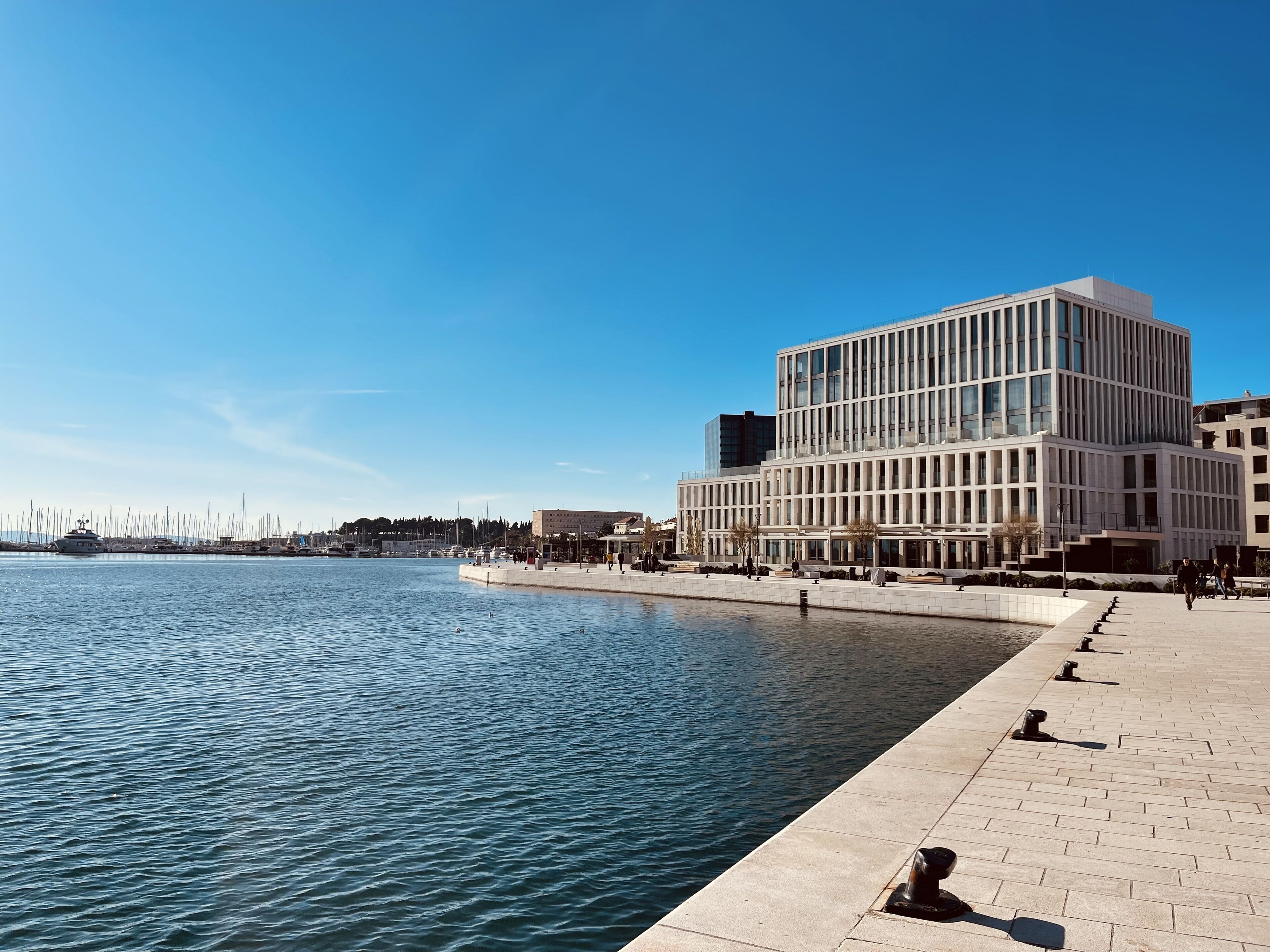 A waterfront building with a modern design, a paved walkway, and a clear blue sky.