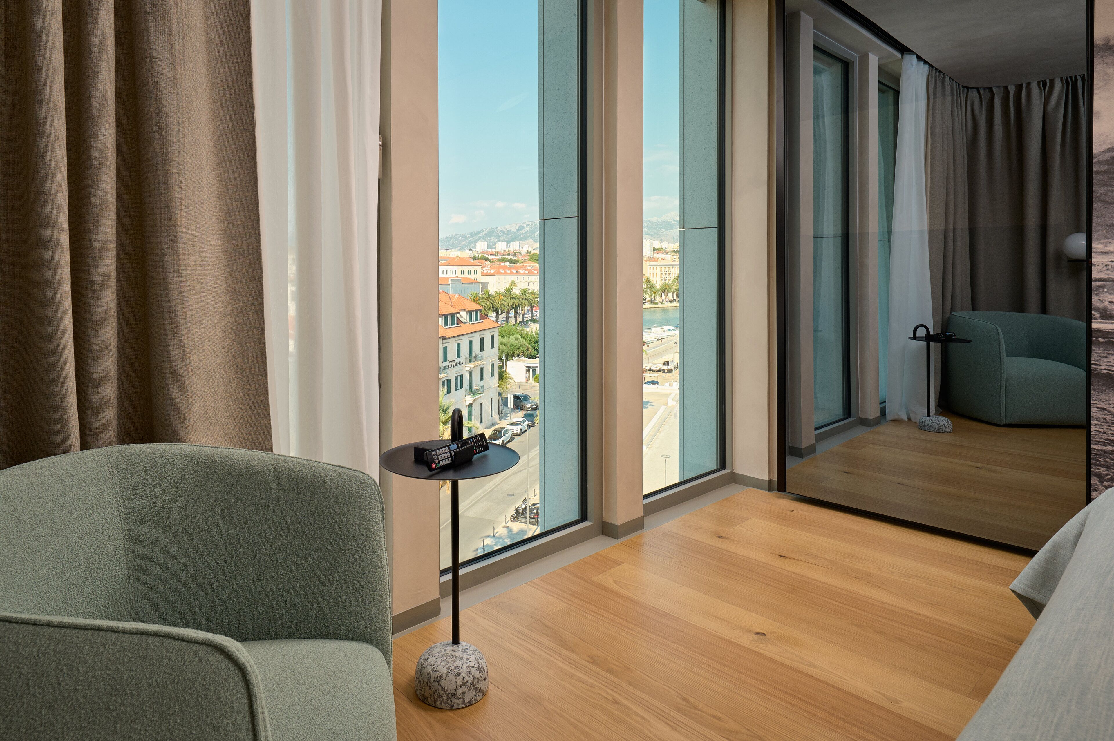 Ein modernes Hotelzimmer mit großem Fenster, einem grünen Sessel, einem kleinen runden Tisch und Blick auf die Stadtlandschaft.