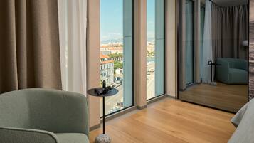 Ein modernes Hotelzimmer mit großem Fenster, einem grünen Sessel, einem kleinen runden Tisch und Blick auf die Stadtlandschaft.
