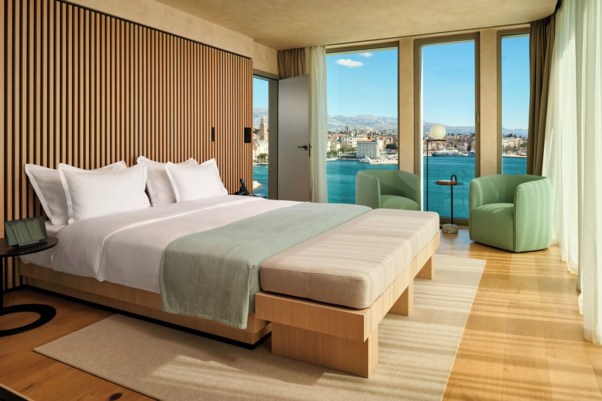 A modern hotel room with a large bed, a view of the sea, and a cityscape.