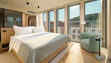 Una habitación de hotel moderna con una cama grande, un espacio de descanso y ventanas amplias con vistas a la ciudad.