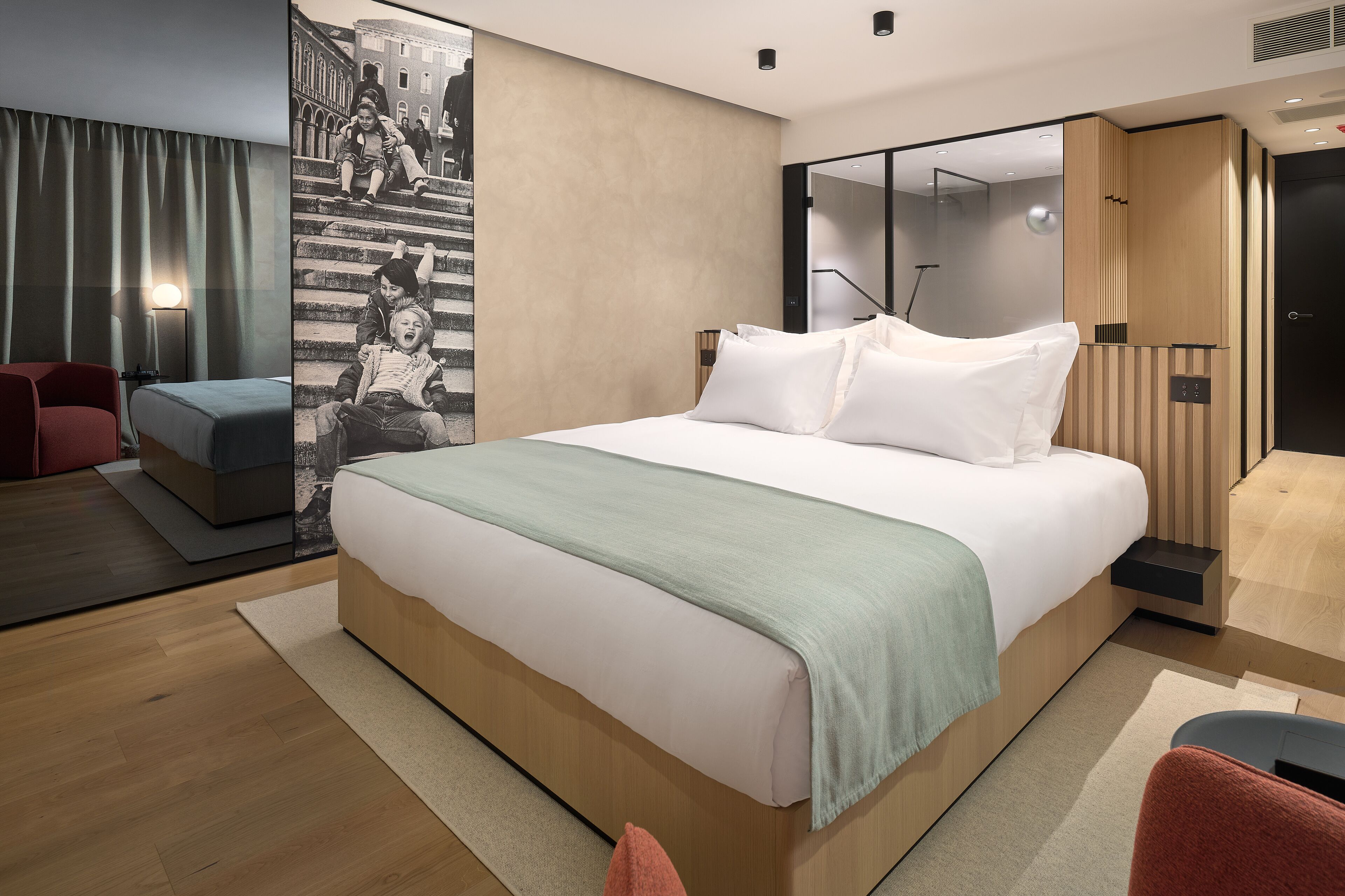 Una habitación de hotel moderna con una cama grande, una zona de estar con una silla roja, un espejo amplio y un televisor montado en la pared.