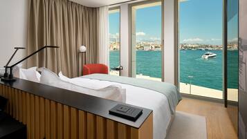 Una habitación de hotel con cama, escritorio con lámpara, una silla roja y un gran ventanal con vista al mar y barcos.