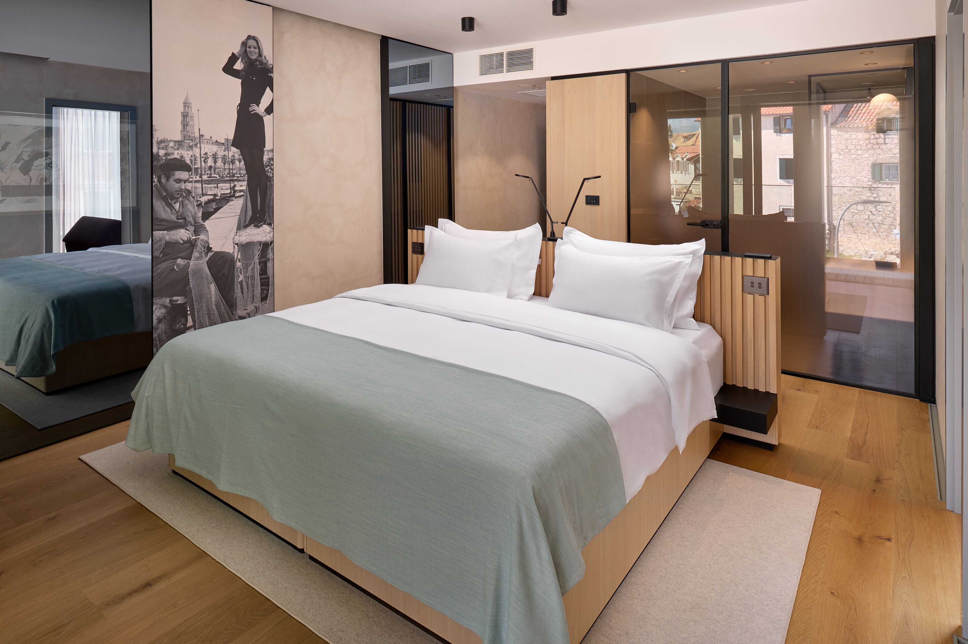 A modern hotel room with a large bed, a smaller bed, a large window, and a view of the city.