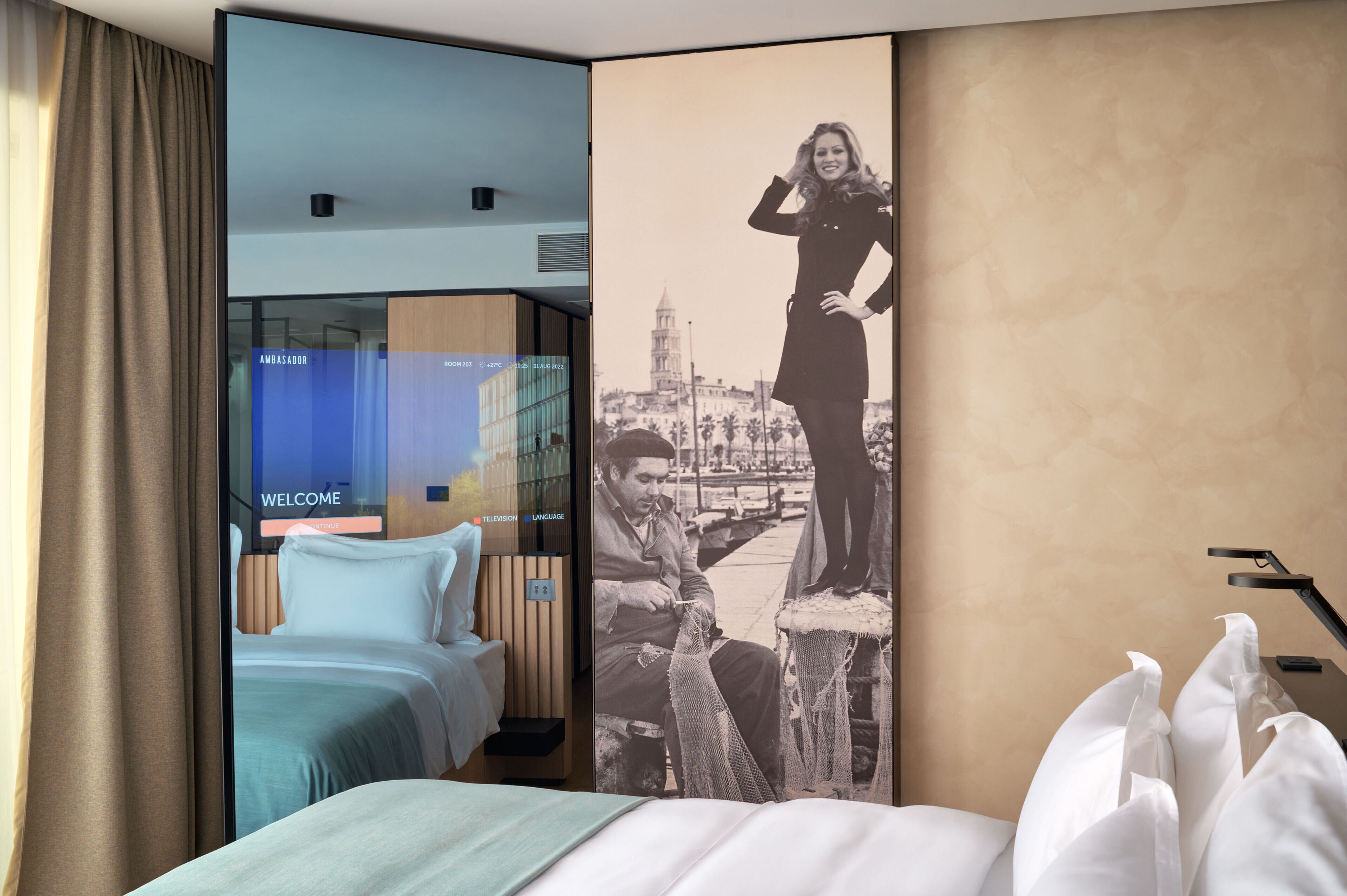 A hotel room with a bed, a TV screen displaying a welcome message, and a large poster of a cityscape.