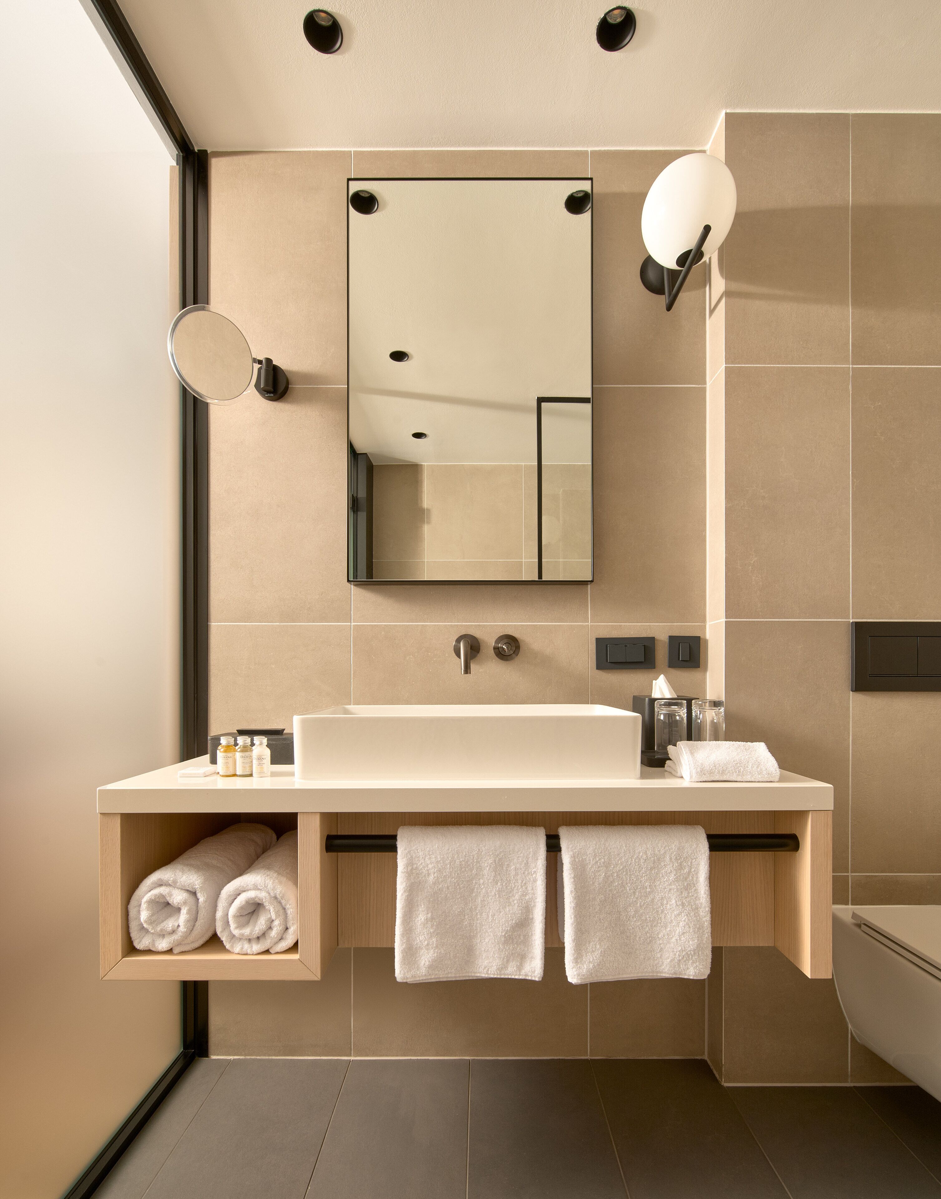 A modern bathroom with a large mirror, a sink, and a shower. There are towels and toiletries on the counter.