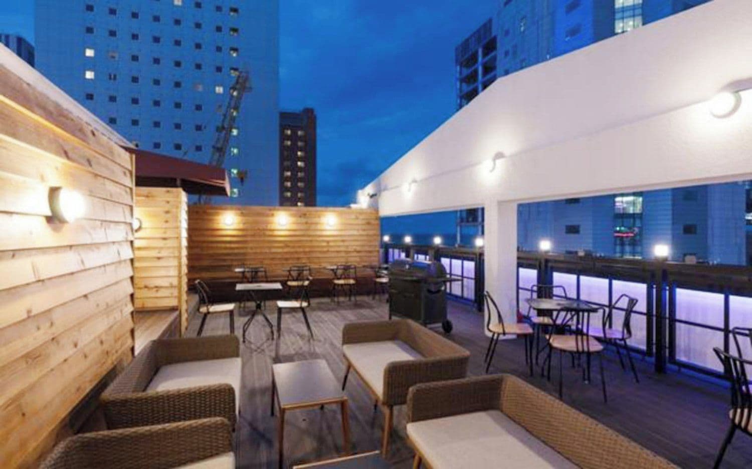 An outdoor seating area with tables and chairs, a wooden wall, and a view of the cityscape at dusk.