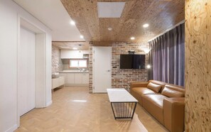 A modern living room with a leather sofa, a coffee table, and a kitchen area with a brick wall and a flat-screen TV. - ARK BLUE HOTEL (Busan)