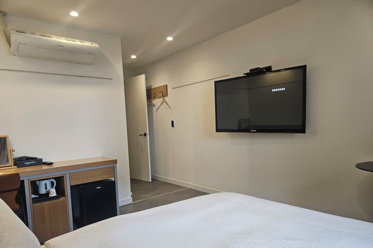 A hotel room with a bed, a TV mounted on the wall, a desk with a kettle, and a door leading to another room.