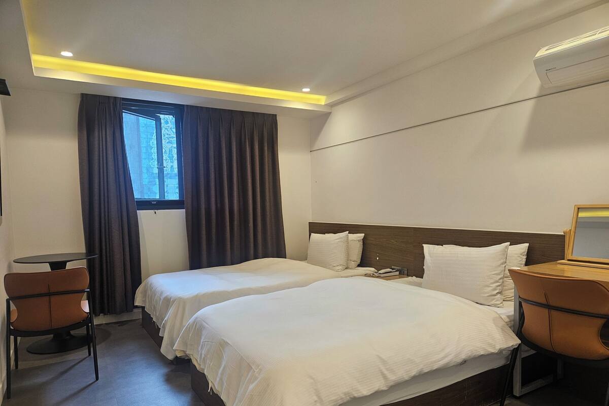 A hotel room with two beds, a small table, chairs, a window with curtains, and an air conditioning unit.