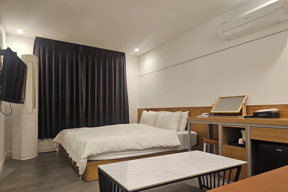 A hotel room with a bed, a desk, a chair, a TV, and an air conditioning unit.