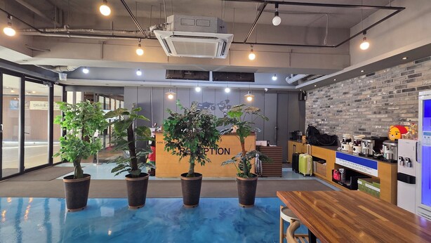 Un moderno vestíbulo de hotel con mostrador de recepción, plantas en macetas, zona de piscina azul y máquina expendedora.