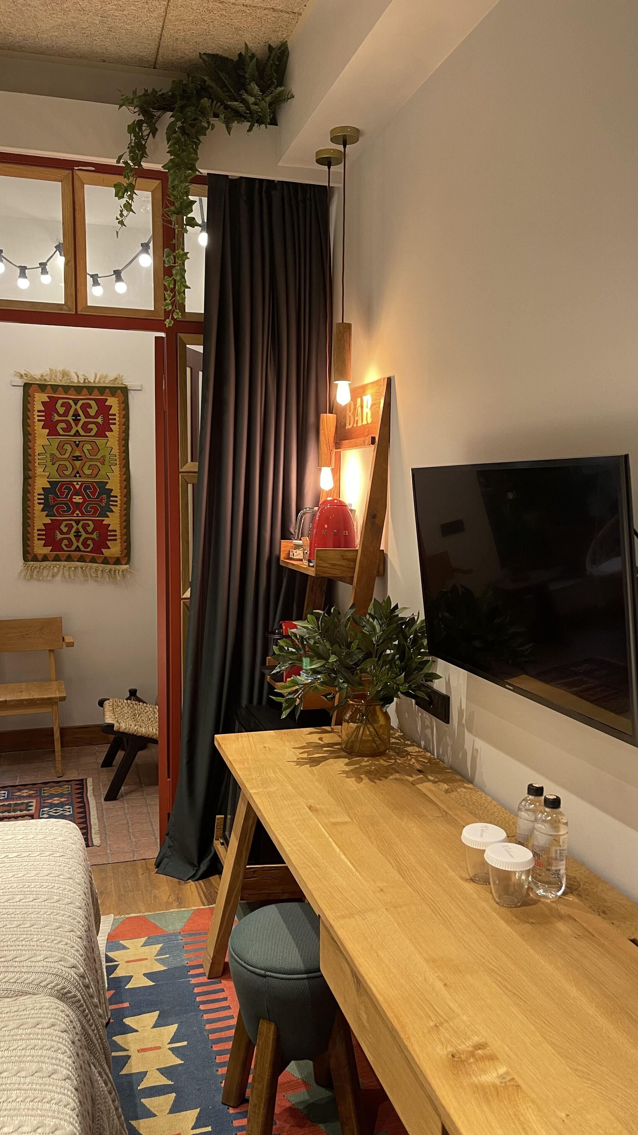 A cozy room with a wooden table, a TV, a window with curtains, a plant, and a wall-mounted shelf with a kettle.