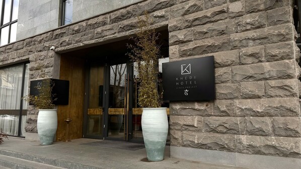 A hotel entrance with stone facade, large potted plants, and signage for 'KHEDI HOTEL GINZA PROJECT'.
