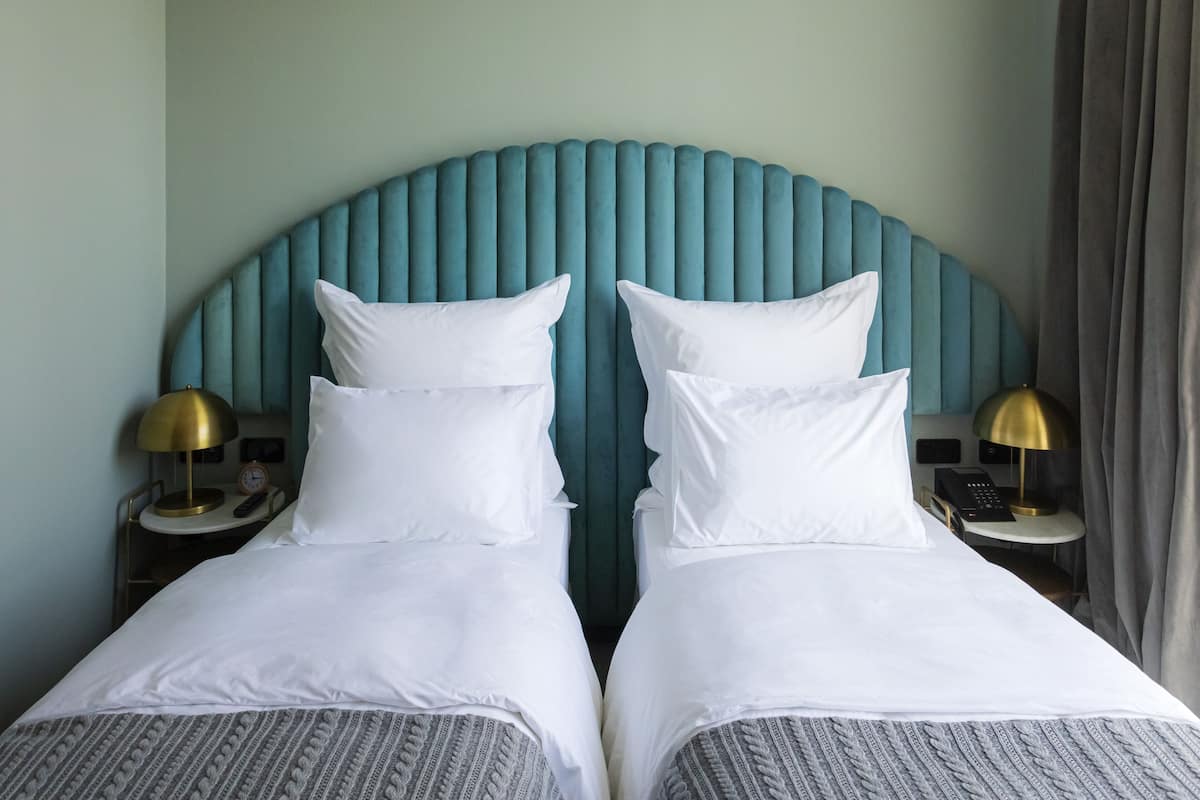 Une chambre d’hôtel avec deux lits, une tête de lit de couleur turquoise, du linge de lit blanc et des lampes de chevet.