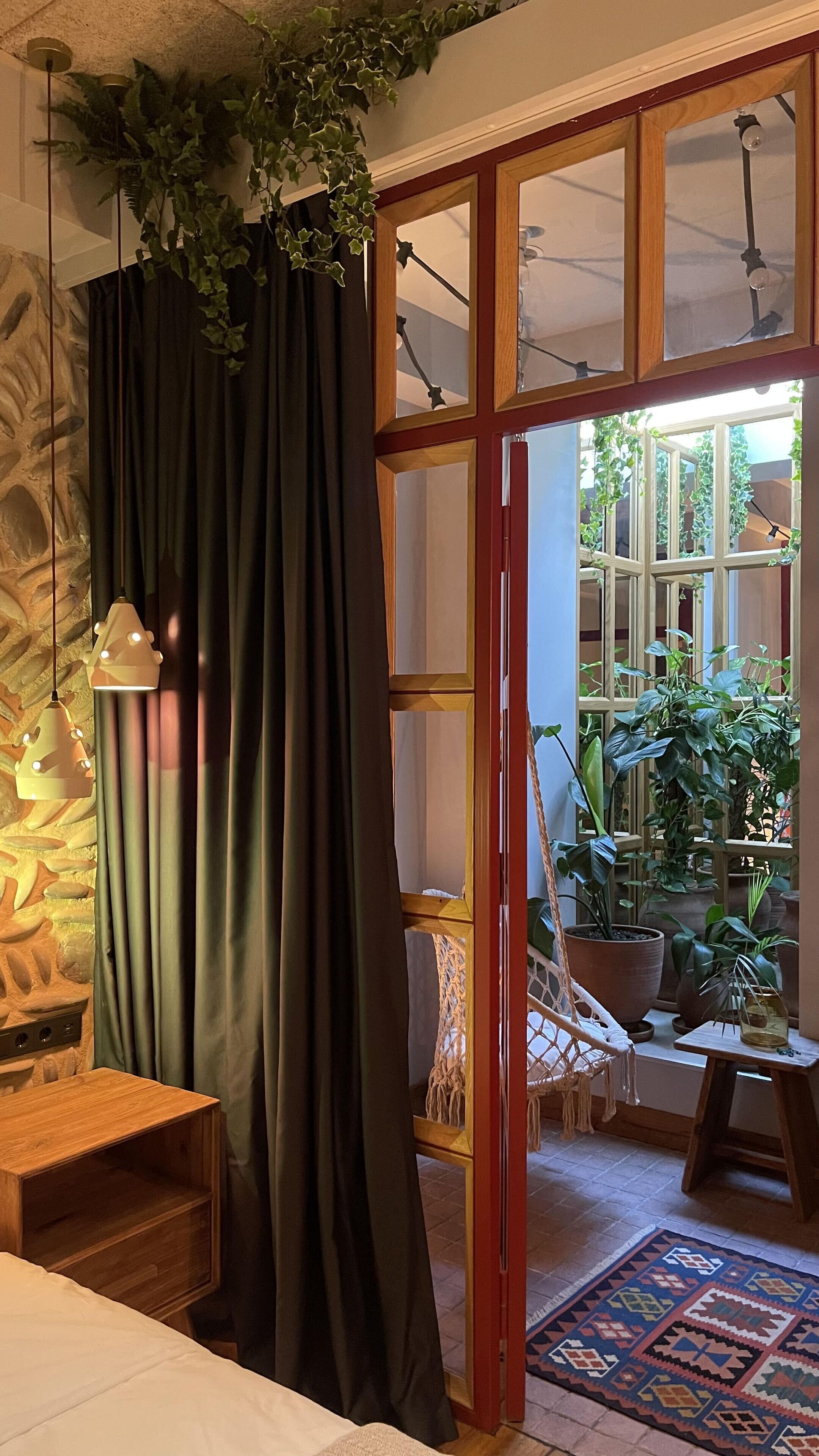 A room with a bed, a wooden nightstand, a macramé chair, a macramé hanging chair, a large window with a view of greenery, and a patterned rug.