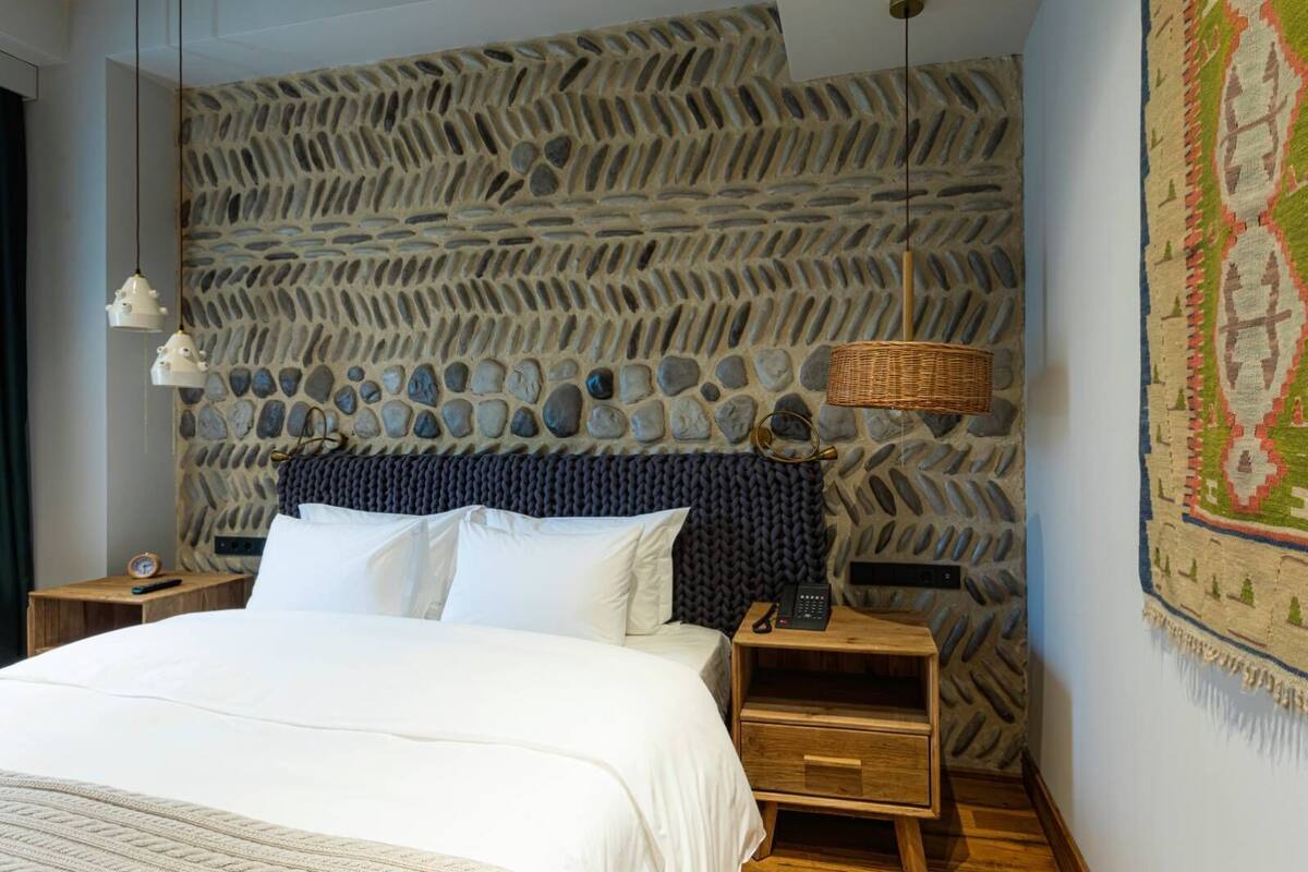 Une chambre à coucher avec une tête de lit à motifs de pierre, un lit avec du linge de lit blanc, une table de chevet avec une lampe et une décoration murale tissée.